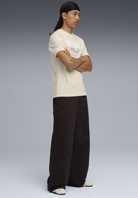 Beige bomulds t-shirt med trykt logo, parret med brune bukser med brede ben. Fodtøj i lys farve med sorte accenter.