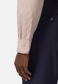 Polsino di camicia di un rosa chiaro con texture e due bottoni, indossato insieme a pantaloni navy a righe sottili. In primo piano, una mano che poggia sul tessuto.