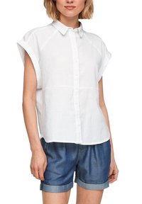 Camicia di lino bianca con maniche corte, chiusura a bottoni e orlo arrotondato. Abbinata a pantaloncini di denim arrotolati per un look casual.