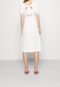 Vestido blanco con estampado floral, mangas abullonadas y un escote en la espalda atado con un lazo. Largo por debajo de la rodilla, combinado con sandalias de tacón rosa.