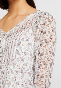Blůza s květinovým vzorem, výstřihem do V, dlouhými rukávy a zapínáním na knoflíky. Vyrobena z lehké, průhledné látky v pastelových odstínech.