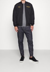 Giubbotto bomber nero con testo ricamato, t-shirt grigia, pantaloni grigi con motivo e sneakers nere con suola bianca. Outfit casual.