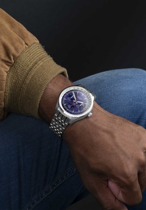 Stainless steel watch with a blue dial, silver accents, and a woven bracelet. Features chronograph sub-dials and a textured casing.