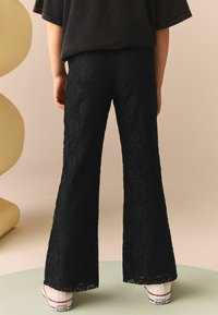 Sorte blonde flared bukser prydet med et blomsterdesign, struktureret overflade og scallop kant, parret med hvide sneakers.