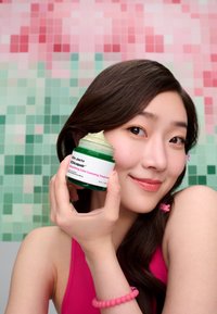 Grønn krukke med en lys grønn krem, merket "Dr. Jart+ Cicapair Soothing Color Correcting Treatment." Bakgrunnen har rosa og grønne fliser.