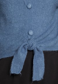 Modrý pletený svetr s měkkou texturou, předními knoflíky a vázaným uzlem v pase.
