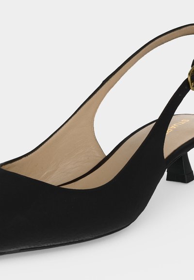 Gros plan sur une chaussure slingback noire à petit talon, intérieur beige et boucle dorée, posée sur un fond uni et clair.