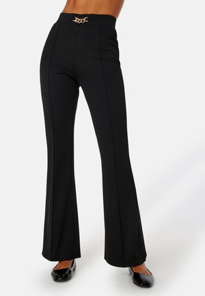 Pantalon noir taille haute évasé avec coutures avant, présentant un détail chaîne dorée à la taille, porté avec des ballerines noires sur fond blanc.