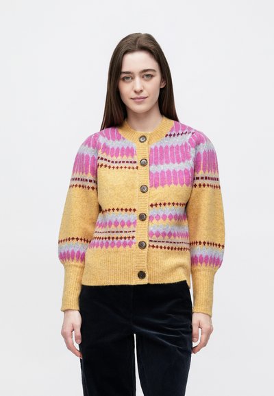 Young woman with straight brown hair wearing a yellow and pink patterned buttoned cardigan and black pants, standing against a white background.