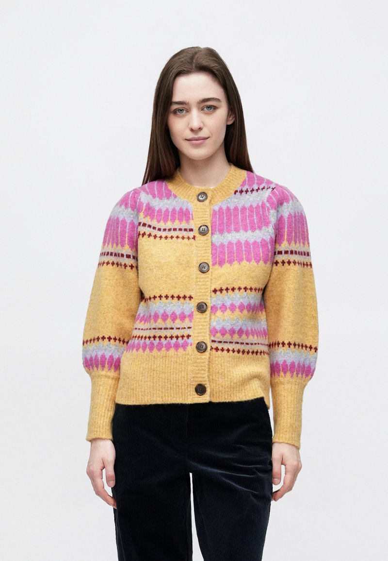 Jauna moteris su tiesiais rudos spalvos plaukais, vilkinti geltonos ir rožinės spalvų raštuotos sagotos megztinis ir juodas kelnes, stovinti prie�š baltą foną.