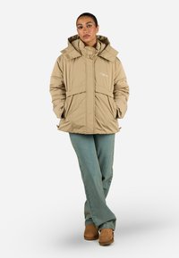 Beige gepolsterte Jacke mit Kapuze, ausgestattet mit einem Frontreißverschluss und zwei Seitentaschen. Der Stoff hat eine glatte Textur und ein dezentes Logo-Detail.