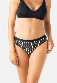 Femme portant un bralette noir et un sous-vêtement à motifs noir et blanc, se tenant debout avec les bras détendus le long du corps sur un fond blanc.