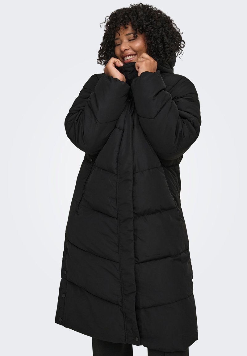 Musta untuvatakki, pitkä ja ylisuurikokoinen malli, jossa tikatut osiot, korkea kaulus ja sivutaskut. Pehmeä, sileä rakenne.