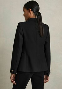 Černý texturovaný blazer s strukturálním designem, zlatými knoflíkovými akcenty a ušitým střihem. Zadní pohled s čistou siluetou.