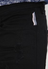 Jeans de mezclilla negra con un detalle desgastado en el muslo y una pequeña etiqueta de marca blanca y roja cerca del bolsillo.