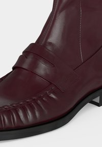 Gros plan d'une chaussure habillée en cuir bordeaux foncé avec un détail de bride, des coutures visibles, et une semelle et un talon noirs.