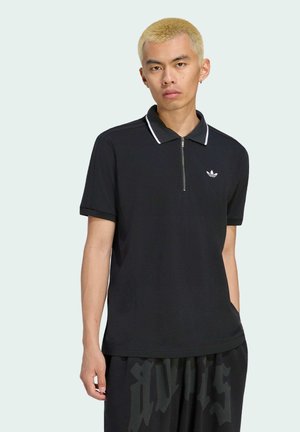 Jeune homme aux cheveux blonds courts portant un polo noir zippé et un pantalon noir avec des lettres foncées sur un fond clair uni.