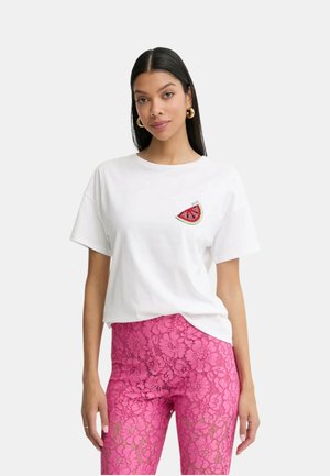 Vrouw met lang zwart haar die een wit T-shirt draagt met een watermeloenpatch en hooggetailleerde roze kanten broek, staand tegen een effen achtergrond.