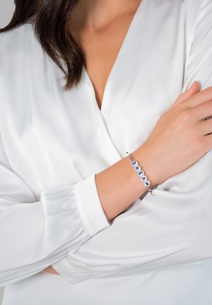 Perlenarmband mit schwarz-weißen Buchstaben, die "DREAM" spelllen. Das Band ist elastisch und wird von einer glatten, weißen Bluse ergänzt.