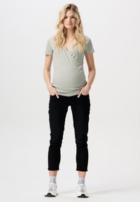 Top de maternité en tissu vert clair, avec un col en V et des manches courtes, associé à un pantalon slim noir et des baskets blanches.