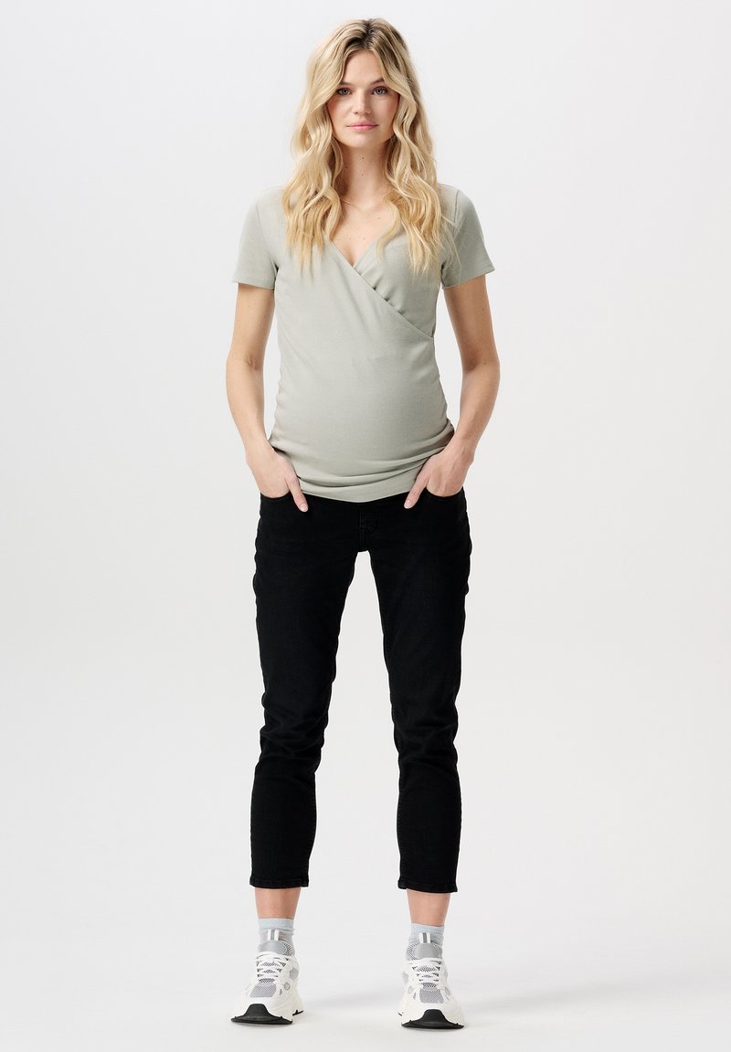 Top de maternité en tissu vert clair, avec un col en V et des manches courtes, associé à un pantalon slim noir et des baskets blanches.