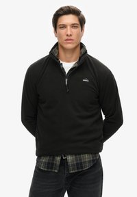 Pullover in pile nero con colletto zip e camicia a scacchi sottostante. Il pullover ha una texture liscia e un logo discreto sul petto.