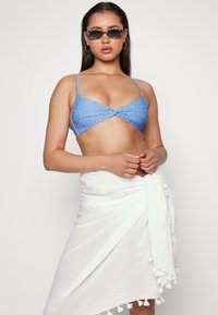 Top de biquíni azul com frente atada e alças finas, combinado com uma fita branca com detalhes em pompons na barra.