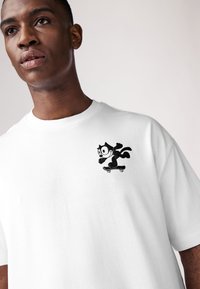 T-shirt en coton blanc avec un graphisme noir représentant un chat sur un skateboard imprimé sur le côté gauche de la poitrine. Col rond et coupe décontractée.