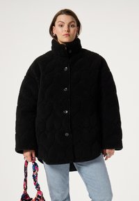 Zwarte fleecejack met een gewatteerde textuur, hoge kraag en knoopsluiting, gecombineerd met een multikleurige bloemenhandtas.