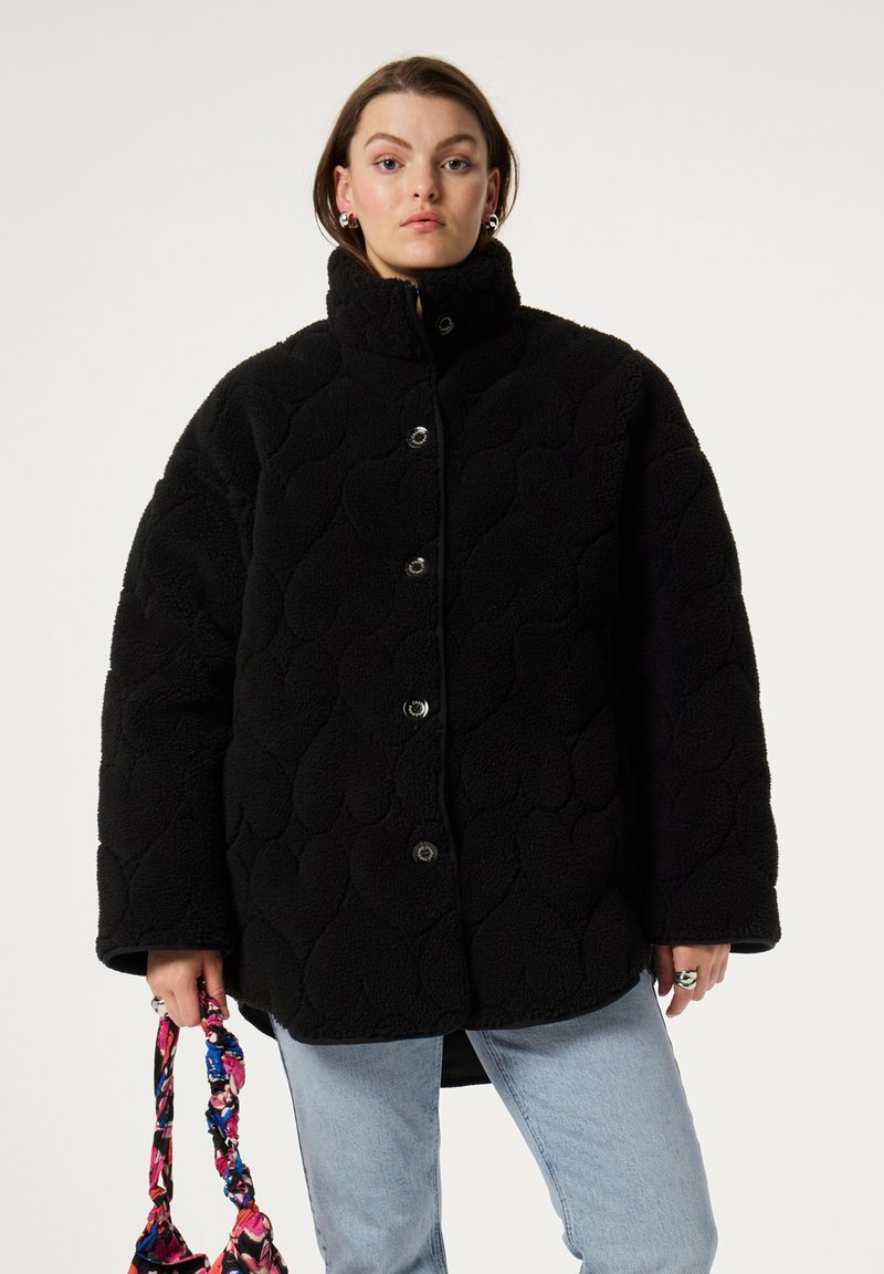 Zwarte fleecejack met een gewatteerde textuur, hoge kraag en knoopsluiting, gecombineerd met een multikleurige bloemenhandtas.