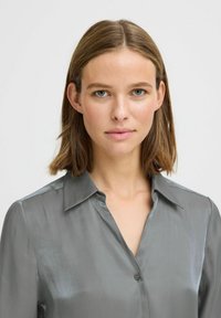 Blusa in satin grigia con colletto, apertura frontale con bottoni e un leggero luccichio. Presenta una linea dritta e una texture liscia. Nessun motivo o accento visibile.