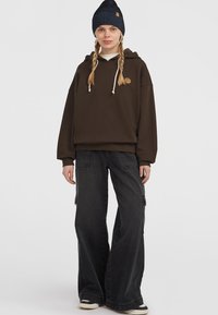 Bruine hoodie van zacht materiaal met een logo-borduursel, gecombineerd met losse zwarte cargo-broeken met zijzakken en een brede pijp.