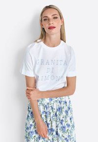 White cotton t-shirt with short sleeves, featuring blue text “GRANITA DI LIMONE.” Worn with a floral blue and white skirt.