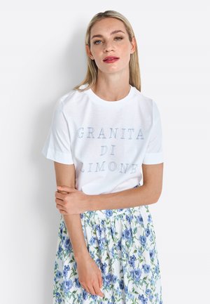 T-shirt en coton blanc à manches courtes, avec du texte bleu "GRANITA DI LIMONE." Porté avec une jupe fleurie bleue et blanche.