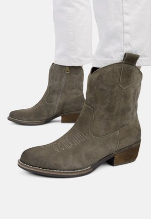 Suede mid-calf brown boots with wooden heel and side zipper, worn with white pants, showing detailed stitching on the toe area.