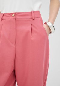 Roze op maat gemaakte broeken met een geplooid ontwerp, zijzakken en een metalen knoopsluiting. Gladde stof met een licht gestructureerde vorm.