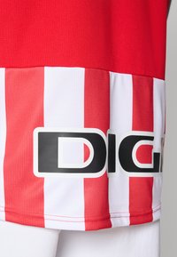 Rot-weißer, senkrecht gestreifter Sporttrikot-Saum mit teilweisem schwarz-weißen "DIGI"-Logo auf dem Stoff über weißen Shorts.
