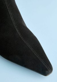 Čierna semišová topánka s špicatou špičkou a viditeľným prešitím pozdĺž priehlavku, s hladkou textúrou, ktorá zdôrazňuje elegantný siluetu a zúžený dizajn.