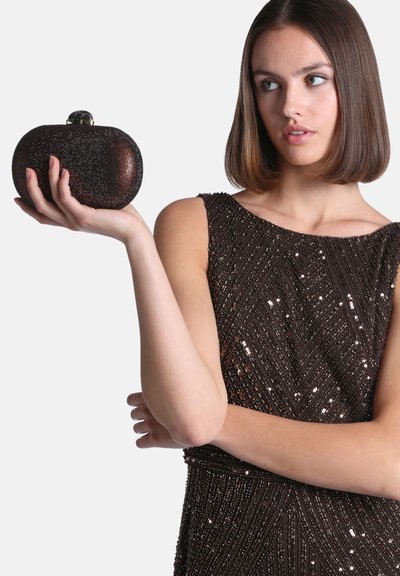 Jeune femme aux cheveux bruns raides en robe noire sans manches pailletée tient une petite pochette noire ovale dans sa main levée.