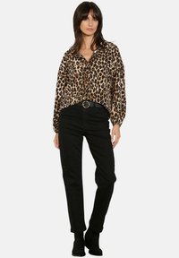 Mujer de pie vistiendo una blusa de manga larga con estampado de leopardo, pantalones negros de cintura alta con cinturón y botas negras hasta el tobillo sobre un fondo blanco.