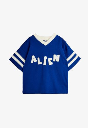 Μπλε πουκάμισο με κοντά μανίκια, λευκή επένδυση V-λαιμού, δύο λευκές ρίγες σε κάθε μανίκι και λευκά γράμματα που σχηματίζουν τη λέξη "ALIEN" στο στήθος.