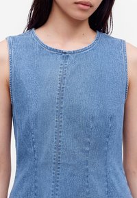 Femme portant un haut en denim bleu clair sans manches avec un col rond, montrant le torse supérieur et le cou, sur fond blanc.