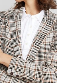 Blazer a cuadros en tonos beige, negro y naranja con un diseño estructurado, solapas en pico y botones en los puños. Camisa blanca de cuello debajo.