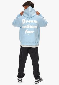 Hellblauer Hoodie mit dem Aufdruck "Träume ohne Angst" in Weiß auf dem Rücken. Mit einer Kapuze mit Kordelzug und geripptem Saum. Kombiniert mit schwarzen Cargo-Hosen.