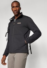 Giacca nera con zip e collo alto, realizzata in tessuto testurizzato. Presenta un patch con logo e cinturini regolabili sulle spalle.