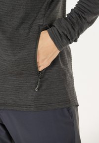 Maglia a maniche lunghe di colore grigio scuro, con tasca con zip e tessuto testurizzato. I polsini sono arricciati e si abbina a pantaloni scuri.