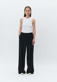 Bela brezrokavnik, oblečen z visokim pasom, ohlapnimi črnimi hlačami in špičastimi bež čevlji. Manekenka stoji z rokama v žepih.