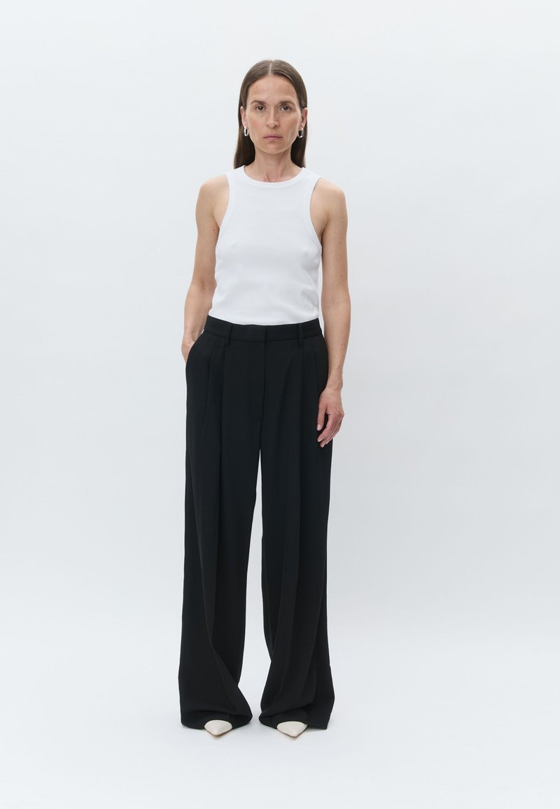 Bela brezrokavnik, oblečen z visokim pasom, ohlapnimi črnimi hlačami in špičastimi bež čevlji. Manekenka stoji z rokama v žepih.