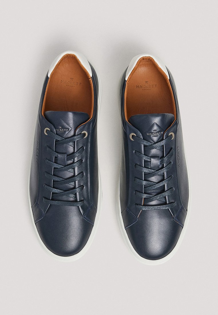 Baskets en cuir bleu marine avec une finition lisse, accents de talon blancs et doublure intérieure brune. Présente des lacets plats et un logo discret.