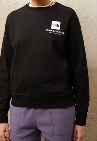 Černá mikina vyrobená z měkké tkaniny, která je zdobena bílým logo ve tvaru čtverce s koordinátami. Kombinována s strukturovanými levandulovými kalhotami s kapsami.